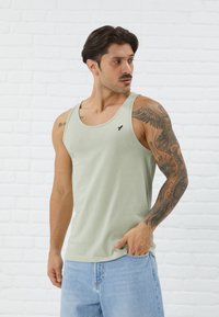 Light green cotton tank top with a crew neckline, featuring a small black bird logo on the chest, paired with light blue denim shorts.
