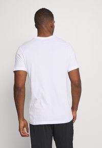 Man sett från bakom iklädd en enkel vit t-shirt med korta Ärmar och svarta byxor mot en ljusgrå bakgrund.