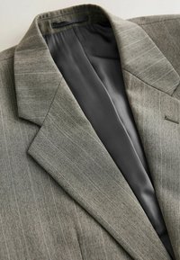 Veste de costume gris à fines rayures avec larges revers en encoche et doublure intérieure grise foncée lisse, pliée à plat sur une surface claire.