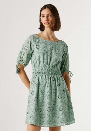 Jonge vrouw met kort bruin haar, gekleed in een lichtgroene jurk met oogjesmotief, pofmouwen en een getailleerde taille, staand tegen een effen achtergrond.