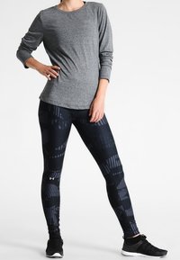 Harmaa pitkähihaisella paidalla, joka on yhdistetty mustiin legginsseihin, joissa on geometrinen kuvio. Legginsseissä on sileä rakenne ja logotesti. Mustat urheilusandaalit.