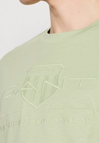 T-shirt vert clair en tissu doux, présentant un logo en relief et un design de blason avec le texte "1949" et "AMERICAN SPORTSWEAR".