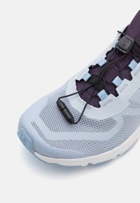 Lyseblå idrettssko med mesh-overdel. Har elastiske lisser, lilla detaljer og strukturert sålemønster. Teksturert design for grep.