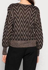 Kobieta ubrana w czarny sweter w metaliczny brązowy wzór w zygzak z bufkami na długich rękawach i czarne spodnie, widziana z tyłu.