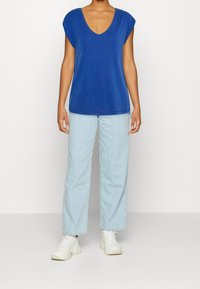 Blue, short-sleeve top with a deep V-neck, paired with light blue wide-leg jeans and white sneakers. Smooth fabric, casual fit.