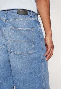 Lyseblå denim jeans har baglommer, et lædermærke i taljen og synlige syninger. Stoffet har en blød tekstur.