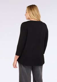 Žena s blond vlasy, oblečená v černém topu s dlouhým rukávem a šedých kalhotách, stojí čelem odvrácena proti jednobarevnému světlému pozadí.
