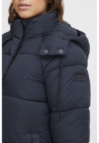 Marineblauwe pufferjas met een geruit patroon, hoge kraag en drukknoopsluiting, voorzien van een logopatch en zijzakken. Gestructureerde stof.