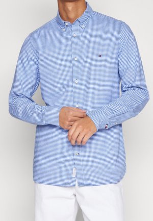 Muž v modro-bílé košili s malým károvaným vzorem a dlouhým rukávem, upravující levý rukáv pravou rukou, má na sobě bílé kalhoty.