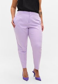 Kvinna klädd i lila avsmalnande byxor och lila högklackade slingback-skor, händerna vilande i fickorna, svart ärmlös topp.