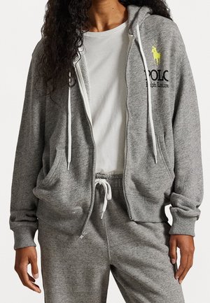 Personne portant un sweat à capuche gris zippé avec un logo jaune Polo Ralph Lauren, un t-shirt blanc et un pantalon de jogging gris assorti avec cordon de serrage.