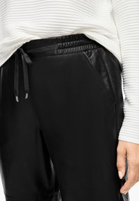Gros plan sur la main d'une personne reposant sur le côté, portant un pantalon en cuir noir avec une taille à cordon et un haut à manches longues côtelé blanc.