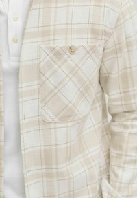 Beež ja valge ruuduline särk, millel on nööbitav rindkere tasku, kantud valge soonikkoes särgi kohal, millel on kolm nööpi.