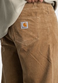 Osoba ubrana w brązowe sztruksowe spodnie z widoczną naszywką Carhartt na tylnej kieszeni oraz beżowy sweter o teksturowanej fakturze.