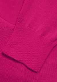 Magenta pletený svetr zblízka, ukazující texturovanou tkaninu s ribkovanými manžetami a hladkým povrchem. Barva je živá a uniformní.