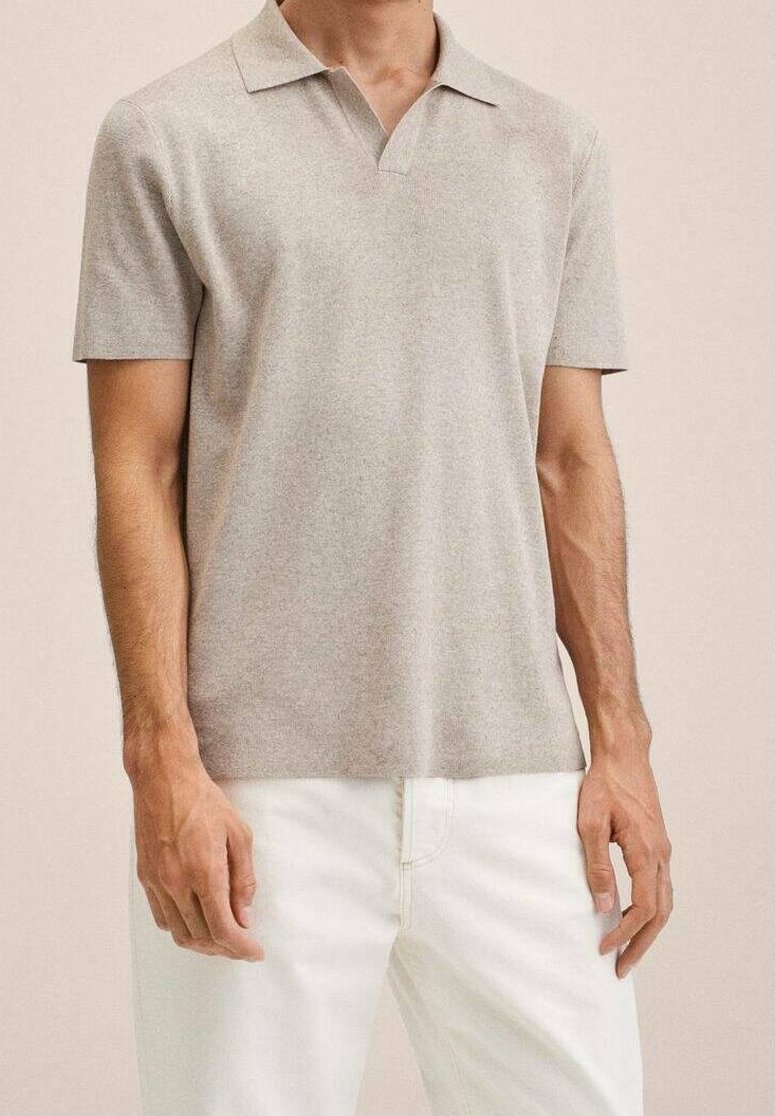 Homme portant un polo beige clair à manches courtes et un pantalon blanc, debout devant un fond neutre.