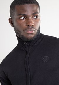 Zwarte trui met ritssluiting, hoge kraag, geribbelde structuur en een klein logo aan de linkerkant van de borst, met een aansluitend ontwerp.