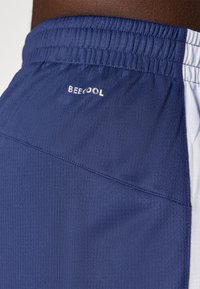 Krupni plan osobe koja nosi plave sportske kratke hlače s elastičnim pojasom i tekstom "BEE COOL" blizu struka.