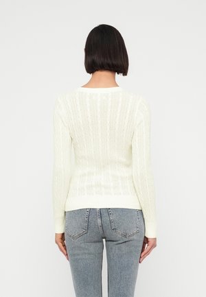 Kvinde med kort, mørkt hår iført en hvid kabelstrikket sweater og lyseblå jeans står med ryggen mod en ensfarvet baggrund.