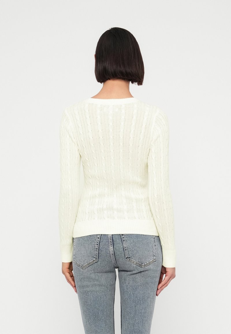 Femme aux cheveux courts et foncés portant un pull en maille torsadée blanc et un jean bleu clair, debout de dos sur un fond uni.