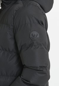 Chaqueta de plumón negra con un tejido suave y brillante, con un diseño acolchado. Parche de logotipo redondo prominente en la parte superior del brazo.