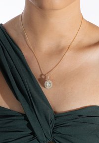 Złoty naszyjnik z łańcuszkiem i kwadratowym wisiorem, w którym centralnie umieszczony jest przezroczysty kamień otoczony mniejszymi kryształkami, noszony na tle ciemnozielonego materiału.