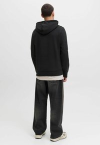 Schwarzer Kapuzenpullover mit Kordelzug, über einem hellen Shirt getragen, kombiniert mit weit geschnittenen dunkelgrauen Jeans und beigen Sneakers.