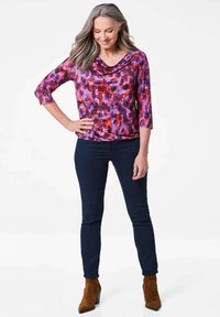 Lila und rosa gemusterte Bluse mit einem Kragen, Dreiviertelärmeln, kombiniert mit dunkelblauen Jeans und braunen Wildleder-Stiefeletten.