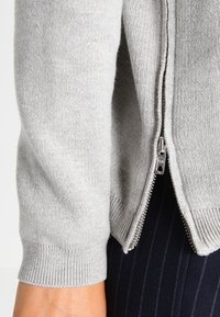 Grå zip-up tröja med ribbade ärmslut, försedda med en metalldragkedja och mjukt tyg. Buren över en randig skjorta, vilket visar lagerdetaljer.