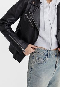 Zwarte leren motorjas met zilveren ritsen, gedragen over een grijze hoodie en lichte spijkerbroek. Voorzien van meerdere met rits afsluitbare zakken en een drukknoopkraag.