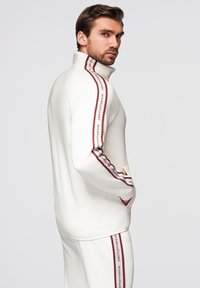 Sudadera blanca con cremallera y cuello alto, con rayas laterales rojas que llevan el texto "performance". Tejido suave y corte relajado.