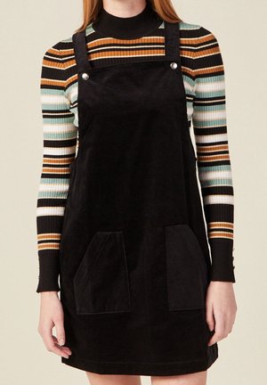 Nainen pukeutuneena mustaan samettiseen halkiamekkoon monivärisen raidallisen pitkähihainen pooloneuleen päällä, seisoo yksiväristä taustaa vasten.