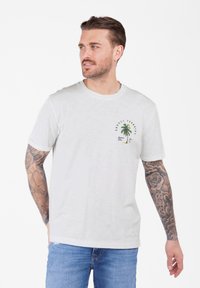Hombre con brazos tatuados que lleva una camiseta blanca con un diseño de palmera y jeans azules, mirando hacia la izquierda contra un fondo blanco.