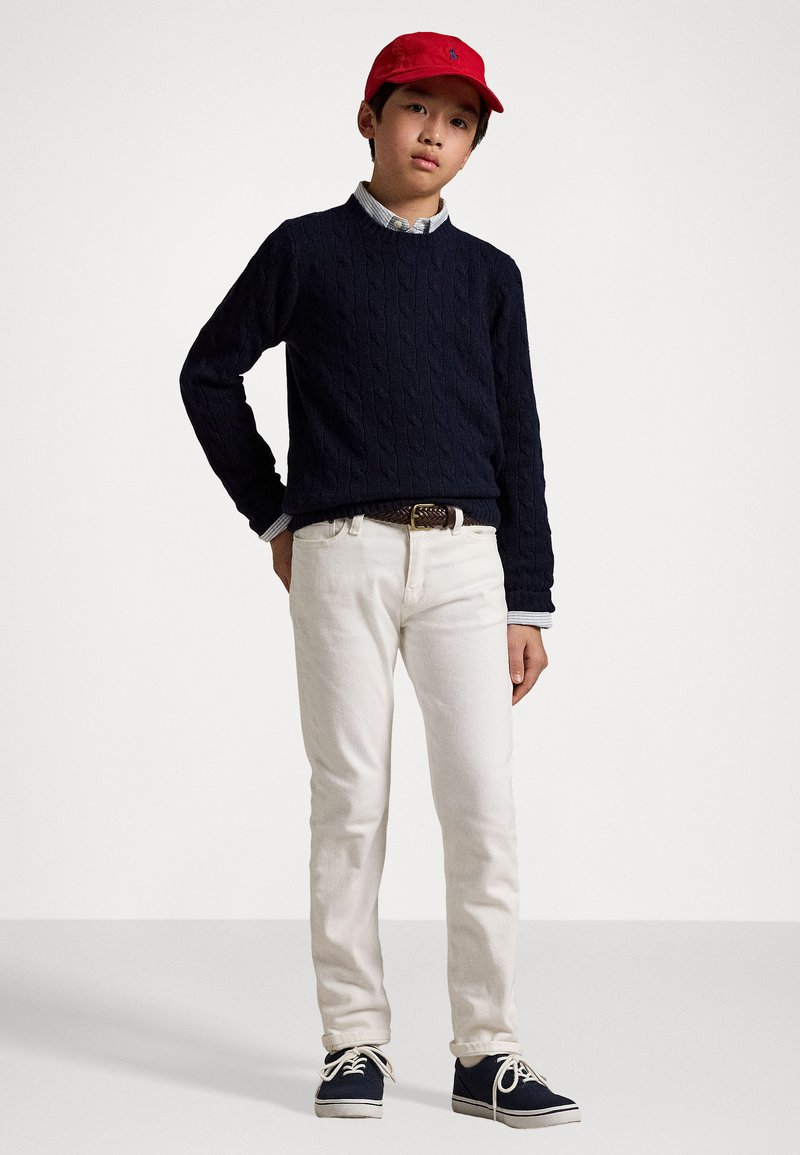 Navy cable-knit sweater, collared shirt beneath, white jeans, red cap, navy sneakers with white soles. Child standing with hands on hips.