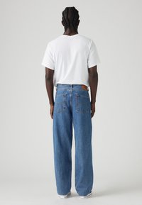 Man med flätat hår iklädd en vit t-shirt och lösa blå jeans, stående vänd bort från kameran mot en ljus, enfärgad bakgrund.