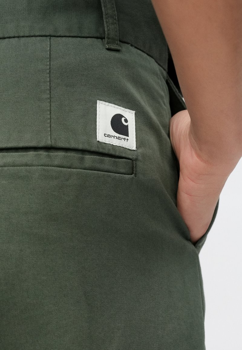 Dunkelgrüne Cargo-Hose mit glatter Textur und einer Gesäßtasche. Auf der Tasche befindet sich ein weißes Markenetikett mit einem schwarzen Logo.