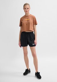 Brun cropped t-shirt med "hummel" logo, siddende sammen med sorte shorts med snøre. Modellen har sorte sportsko på. Enkel atletisk design.
