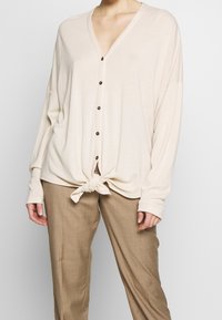 Beige stickad cardigan med V-ringning, långa ärmar och knytfront. Har fem bruna knappar och en avslappnad passform. Matchas med beiga byxor.