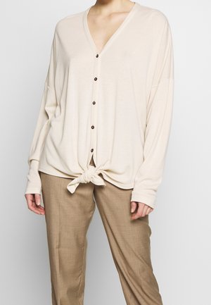 Beige Strickjacke mit V-Ausschnitt, langen Ärmeln und gebundenem Vorderteil. Verfügt über fünf braune Knöpfe und eine lockere Passform. Kombiniert mit beigen Hosen.