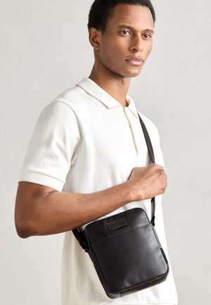 Brown leather crossbody bag with a square shape, stitching detail, and adjustable strap, worn over a cream polo shirt.