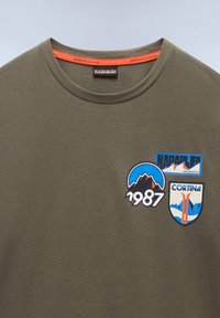 Olivově zelené bavlněné tričko s kulatým výstřihem, na němž se nacházejí modré a černé nášivky hor a potištěné štítky "Napapijri" a "Cortina, 1987."