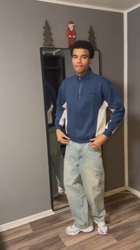 Niebieska bluza na zamek z białymi bocznymi panelami, jasne spodnie jeansowe i białe trampki; stoi przed lustrem na tle szarej ściany.