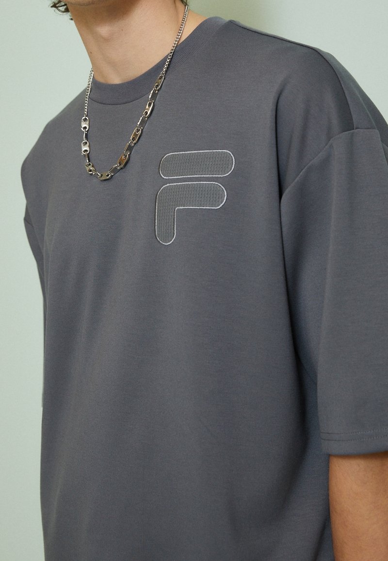 Camiseta gris de manga corta hecha de mezcla de algodón, con un logo texturizado y un escote redondo, acentuada con un collar de cadena plateado.