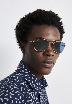 Gold metal frame sunglasses with dark lenses and a varied geometric design. Worn with a navy shirt featuring a white abstract pattern.