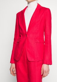 Bright pink linen blazer with a single button closure, notch lapels, two front pockets, and long sleeves, paired with a white turtleneck.