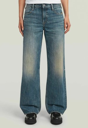 Person trägt ausgewaschene, weite blaue Jeans und schwarze Schuhe und steht vor einem einfarbigen hellen Hintergrund.