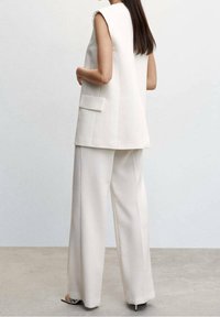 Bílá šitá vesta s bočními kapsami ve spojení s volnými bílými kalhotami. Hladká, strukturovaná látka; minimalistický design.