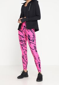 Giacca nera con zip sopra leggings rosa con motivi astratti neri. I leggings hanno una texture elastica, adatta per l'abbigliamento sportivo.