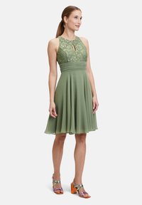 Robe verte avec corsage en dentelle et détail en trou de serrure, jupe en chiffon évasée, longueur genoux, accompagnée de sandales à lanières multicolores.