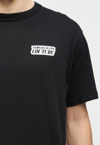 Μαύρο κοντομάνικο μπλουζάκι με λευκό λογότυπο κειμένου "VERSACE JEANS COUTURE" στο αριστερό στήθος.